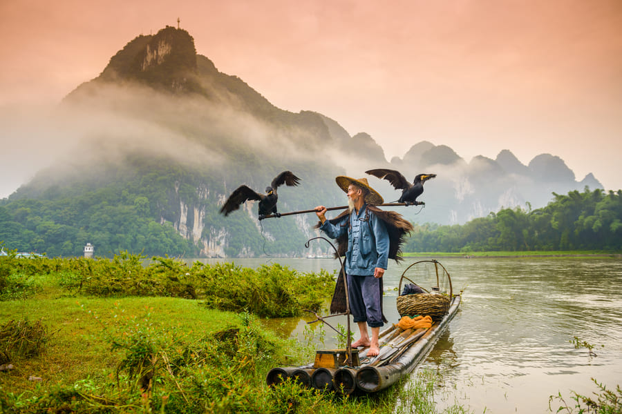 PESCADOR DE CORMORAN EN UNA LANCHA EN EL RIO YOULONG EN YANGSHUO CON MONTAÑAS DE FONDO